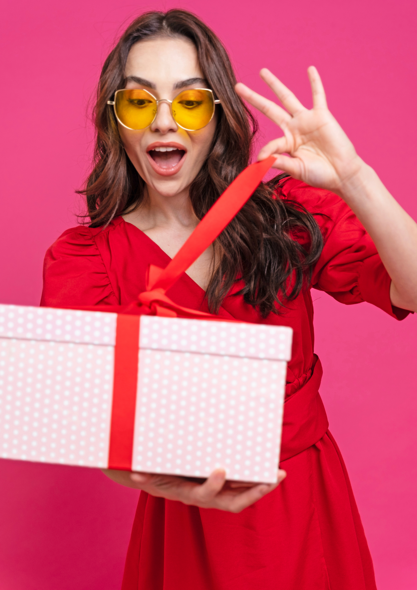 Happy woman in red dress holding a gift box with a red ribbon against a pink background, celebrating with joy representing gift card.